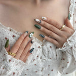 A close-up of a person's hands wearing press-on nails with vibrant green tones and floral detailing with gold accents, holding a floral-patterned fabric.
